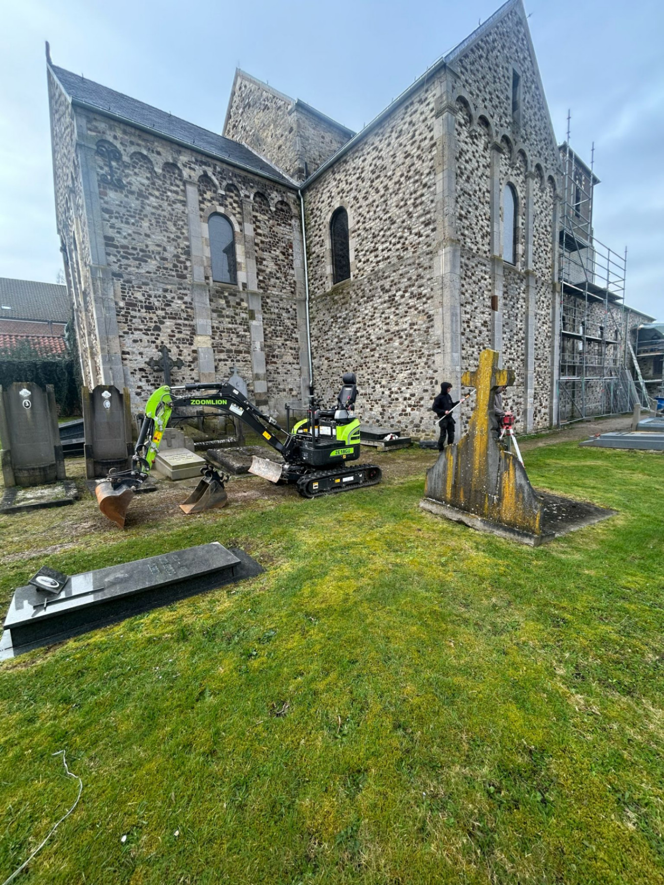 Minigraver van G&S Projecten aan het werk op het kerkhof rond de romaanse kerk van Bierbeek, met archeologen op de achtergrond